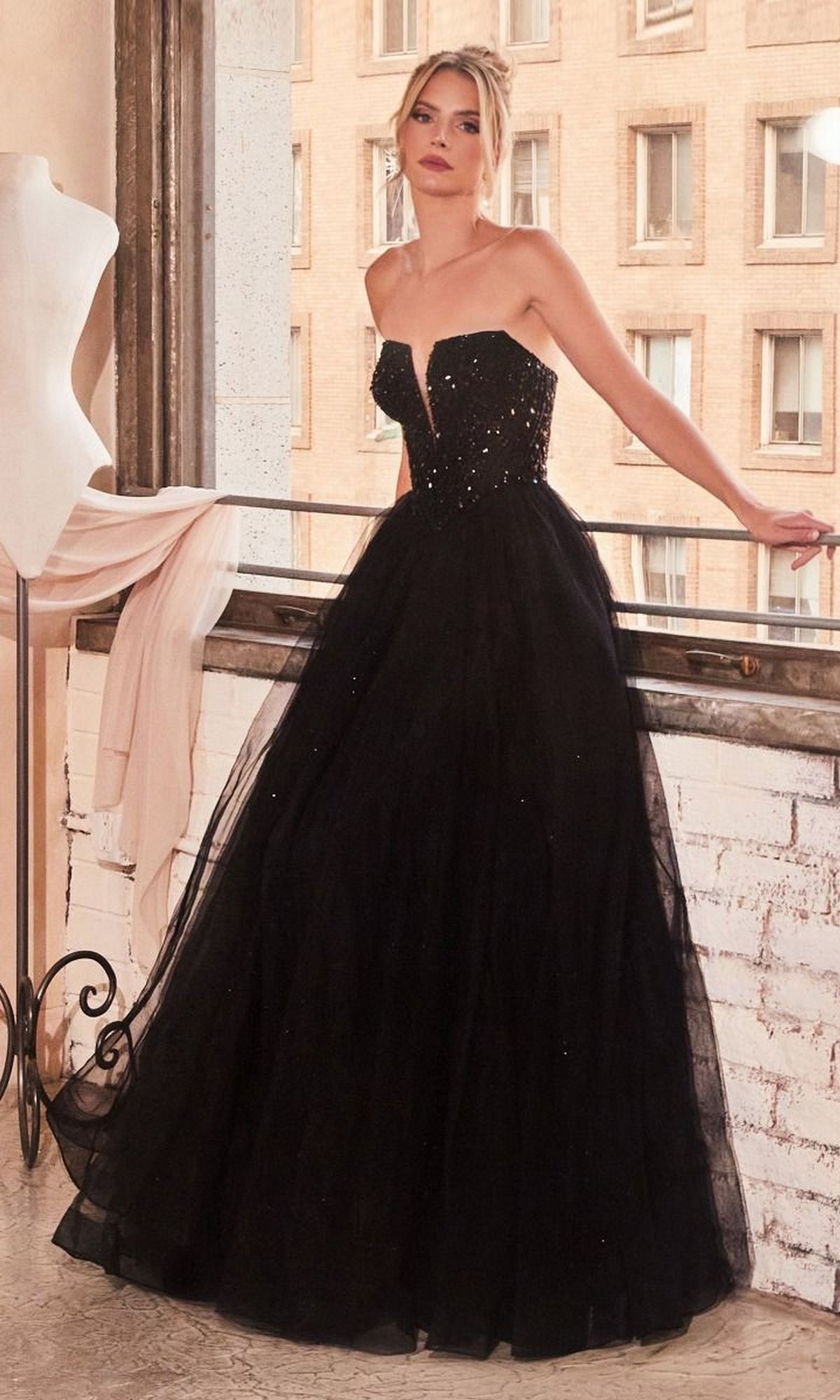 A woman stands by a window in a bright urban room, wearing the Lace-Up Strapless Long Glitter Prom Dress CD0217, featuring a fitted embellished bodice and full tulle skirt.